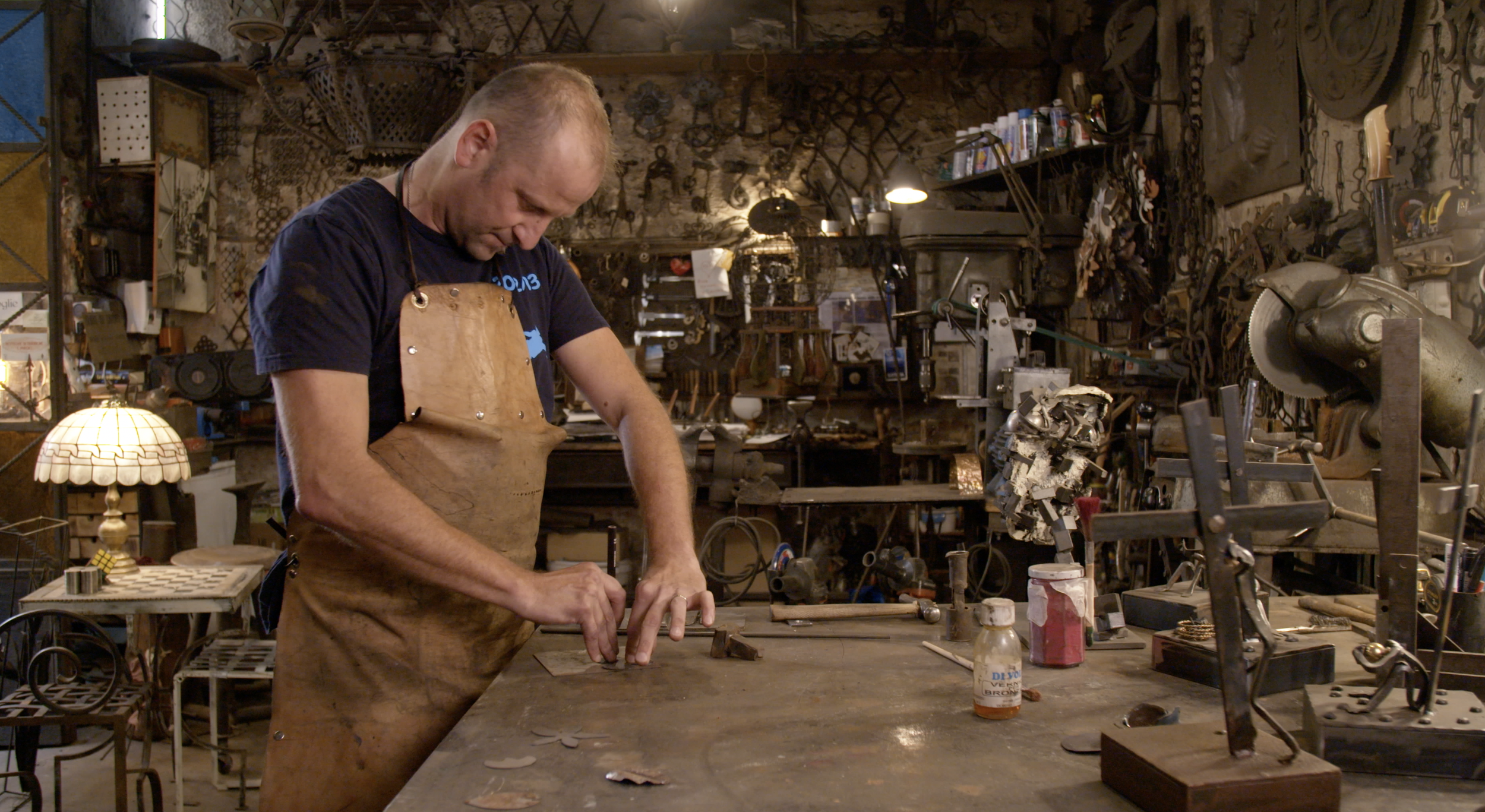 A craftsman in a workshop wearing a leather apron and working on a metal piece at a cluttered workbench. The workshop is filled with tools, metal parts, and shelves. A traditional lamp and various metal sculptures and items are visible in the background.