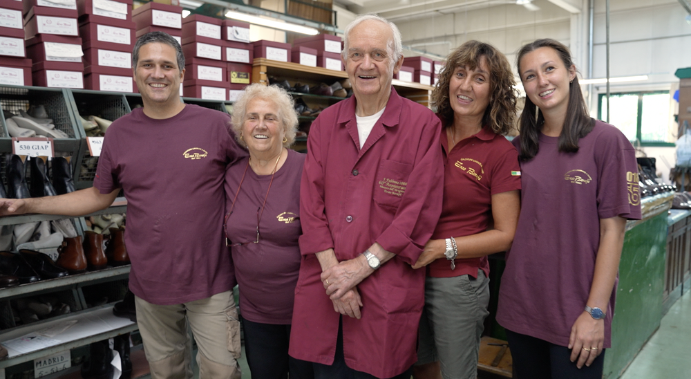 Group of five smiling people wearing matching T-shirts, standing in a shoe store or factory with shelves of shoe boxes and boots in the background.