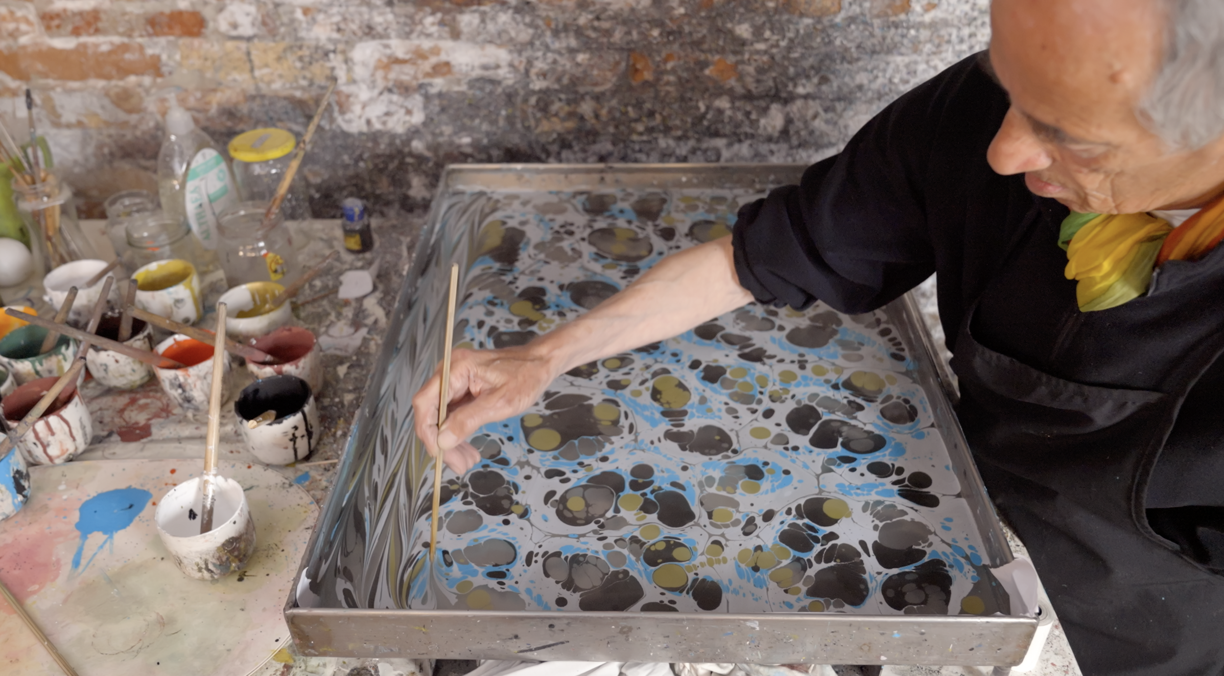 Artist creating marbled paper by using a stick to manipulate paint patterns on water surface, surrounded by cups of paint on a worktable.