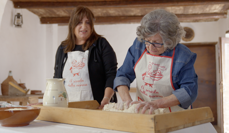 Two women making dough, wearing aprons, in a rustic kitchen setting.