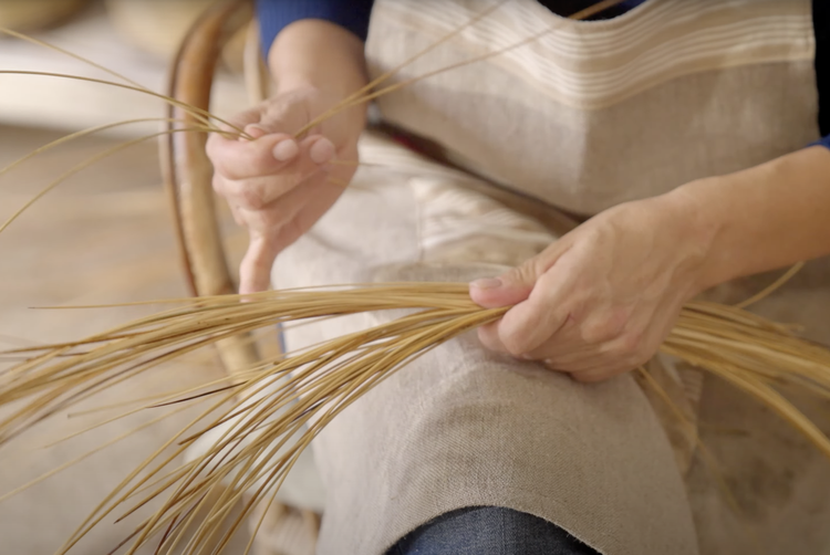 A person weaving with straw, wearing an apron, sitting in a chair.