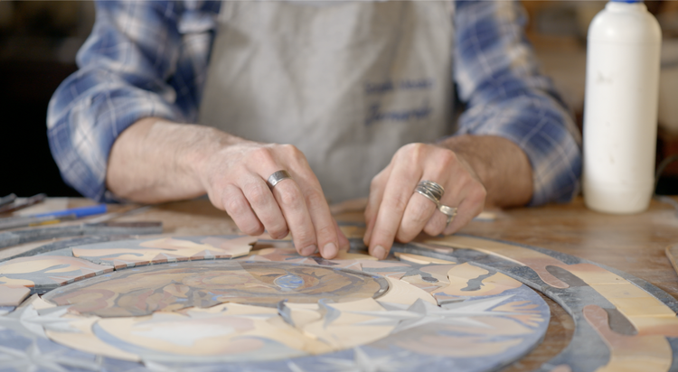 Person arranging tiles on a decorative mosaic, wearing a blue plaid shirt and apron, with various tools and a bottle of glue on the table.
