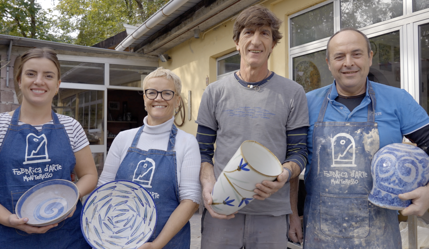 Four people wearing aprons holding handmade pottery outdoors.