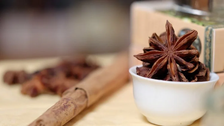 Star anise in a small white bowl on a table with a cinnamon stick and a wooden box nearby.