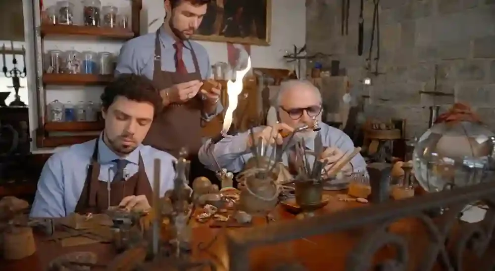 Three artisans working at a jewelry workshop bench surrounded by tools and materials.