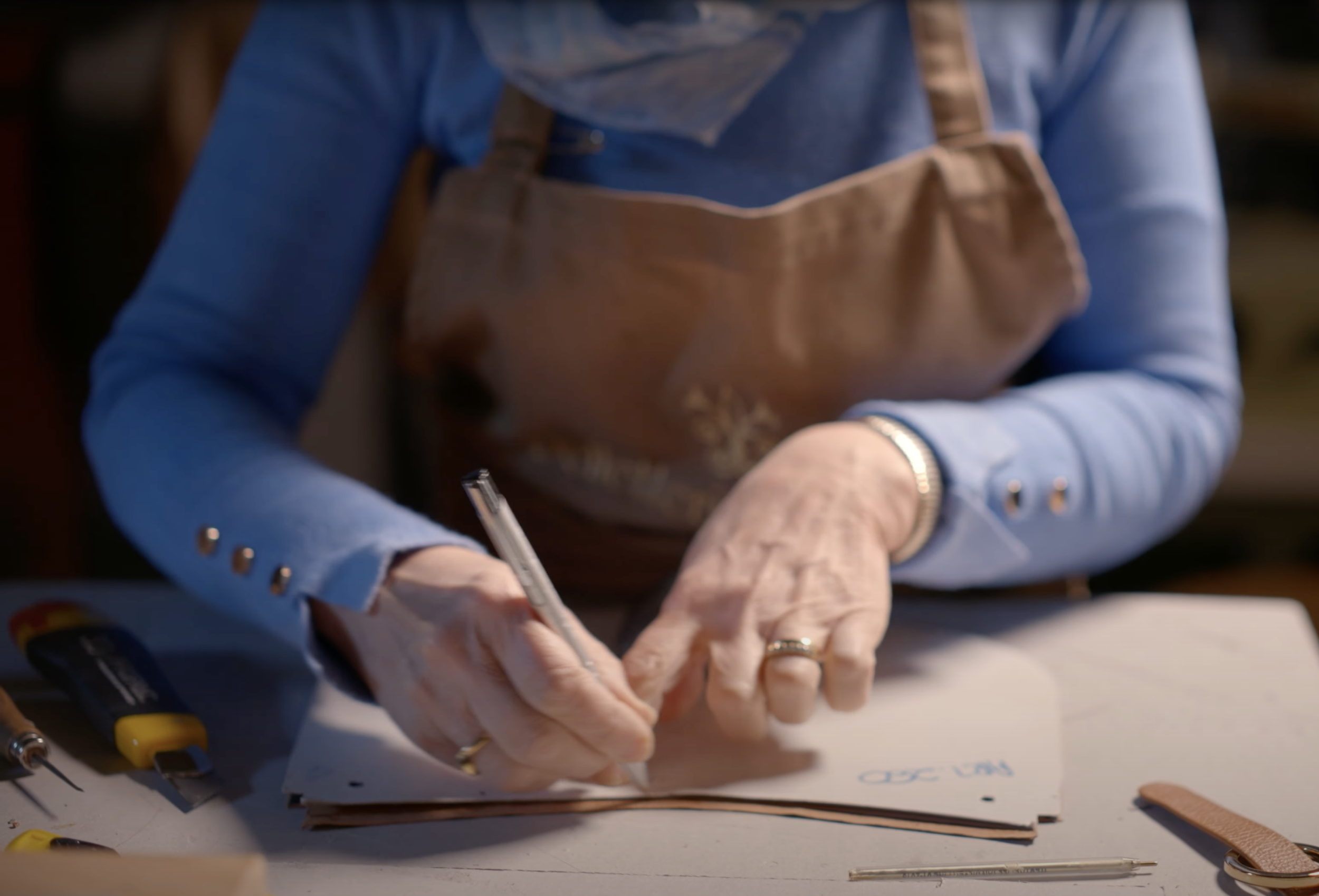 Person wearing a blue shirt and apron, marking leather pieces on a worktable with cutting tools and a pen.