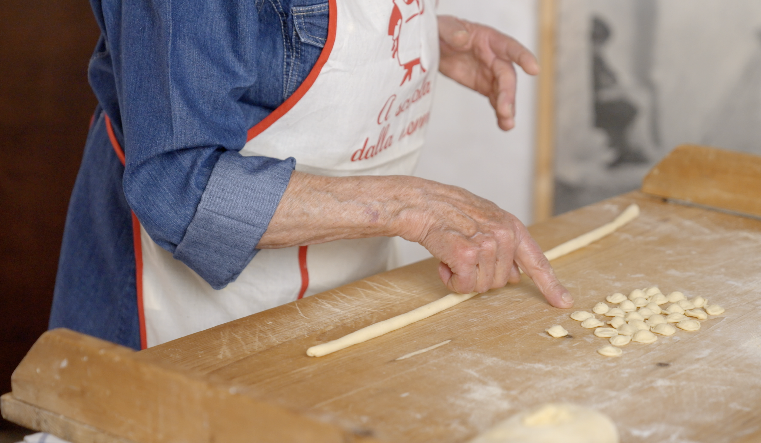 Person in a denim shirt and apron making orecchiette pasta on a wooden surface.