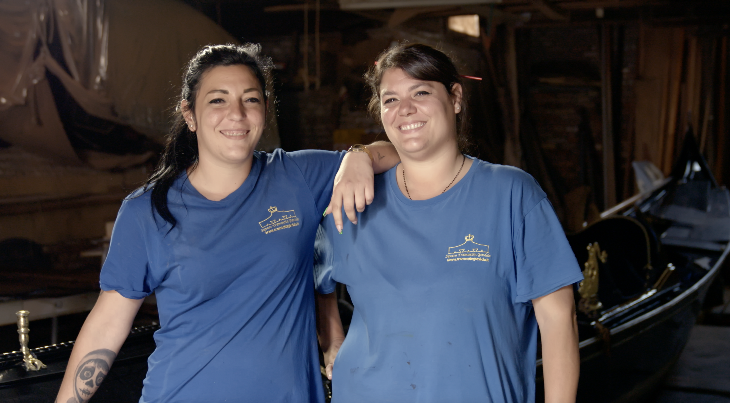 Two women wearing blue shirts, standing in a workshop with a gondola in the background, smiling at the camera.