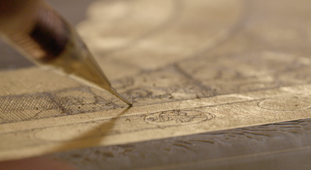Close-up of a pencil tip sketching details on a metallic surface with intricate patterns.