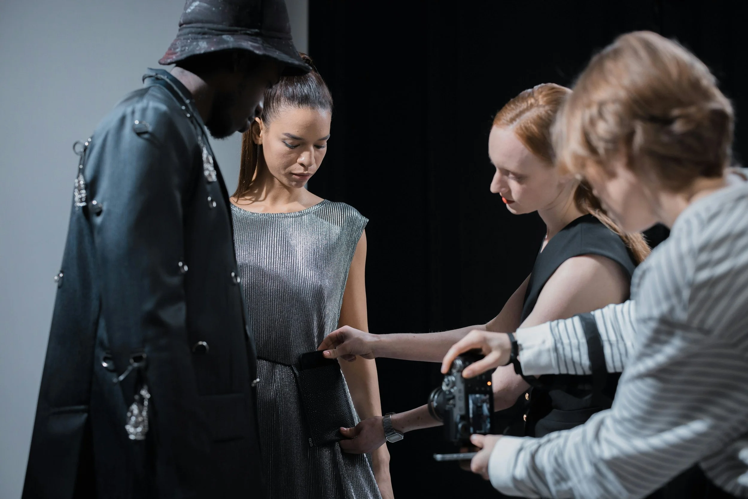 People on a photoshoot set, with a female model in a metallic dress, being adjusted by a stylist, while a photographer is taking pictures.