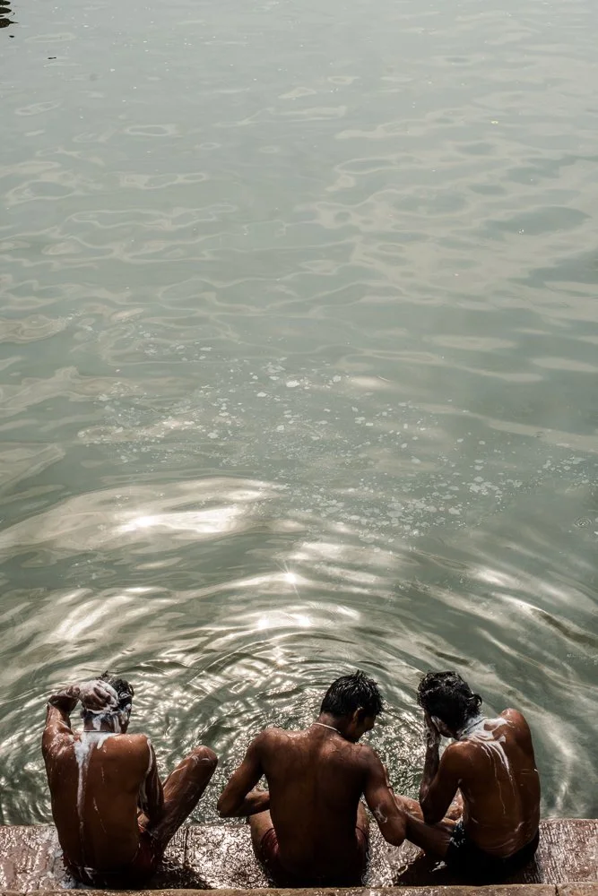 Three young men sitting on a wooden dock at the edge of a body of water, washing their hair with soap and water.