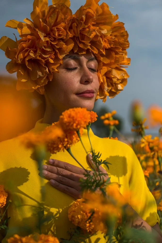 A woman with closed eyes and a peaceful expression, wearing a large orange flower hat and a yellow dress, standing among orange marigold flowers.