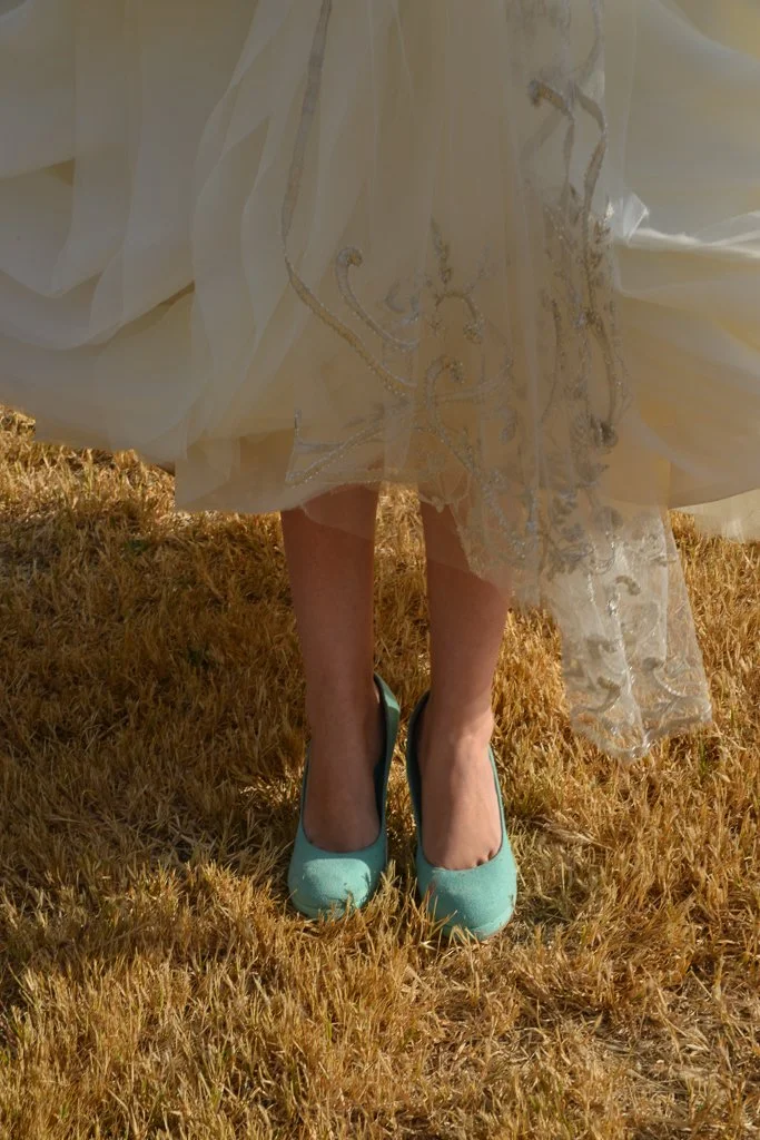 Close-up of a woman wearing teal high heels and a flowing, embroidery dress, standing on dry grass.