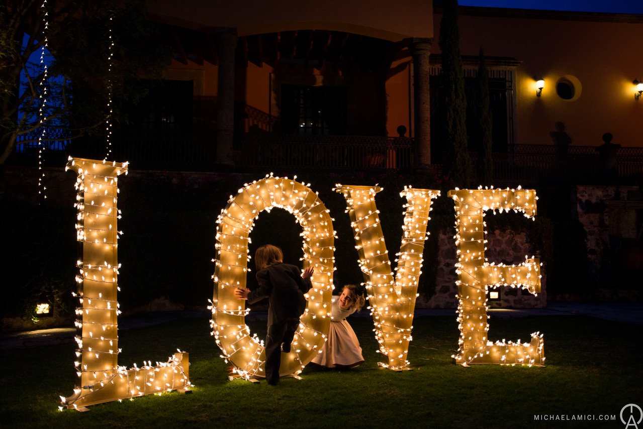 Large illuminated LOVE sign with two children playing in front of it at night. The sign is decorated with small white lights, and the background shows a building with warm lighting.