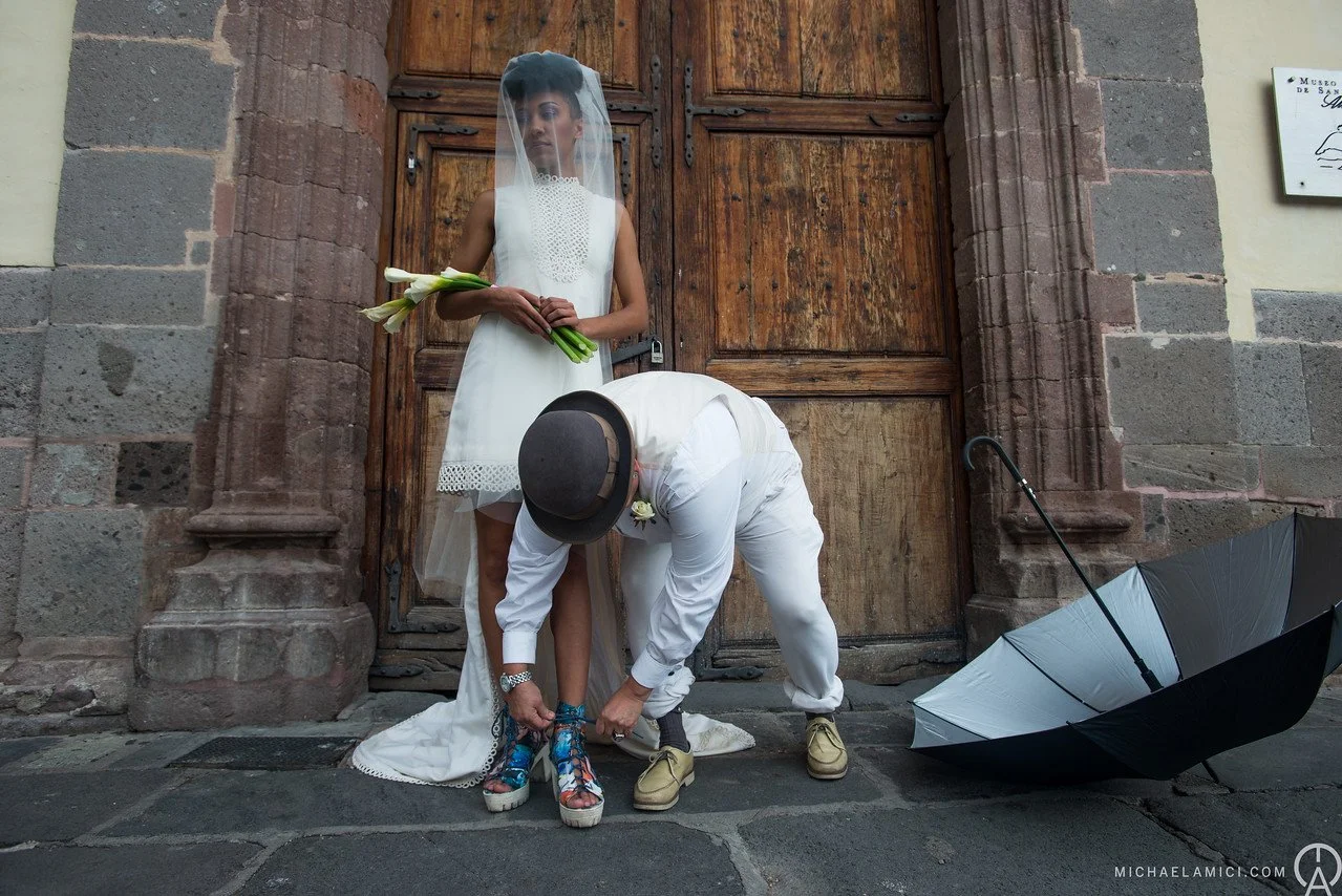 A bride in a white dress and veil stands against a wooden door, holding a bouquet of white flowers. A man in white clothing and a hat is kneeling to tie her colorful shoes. An open black and white umbrella is on the ground beside them.