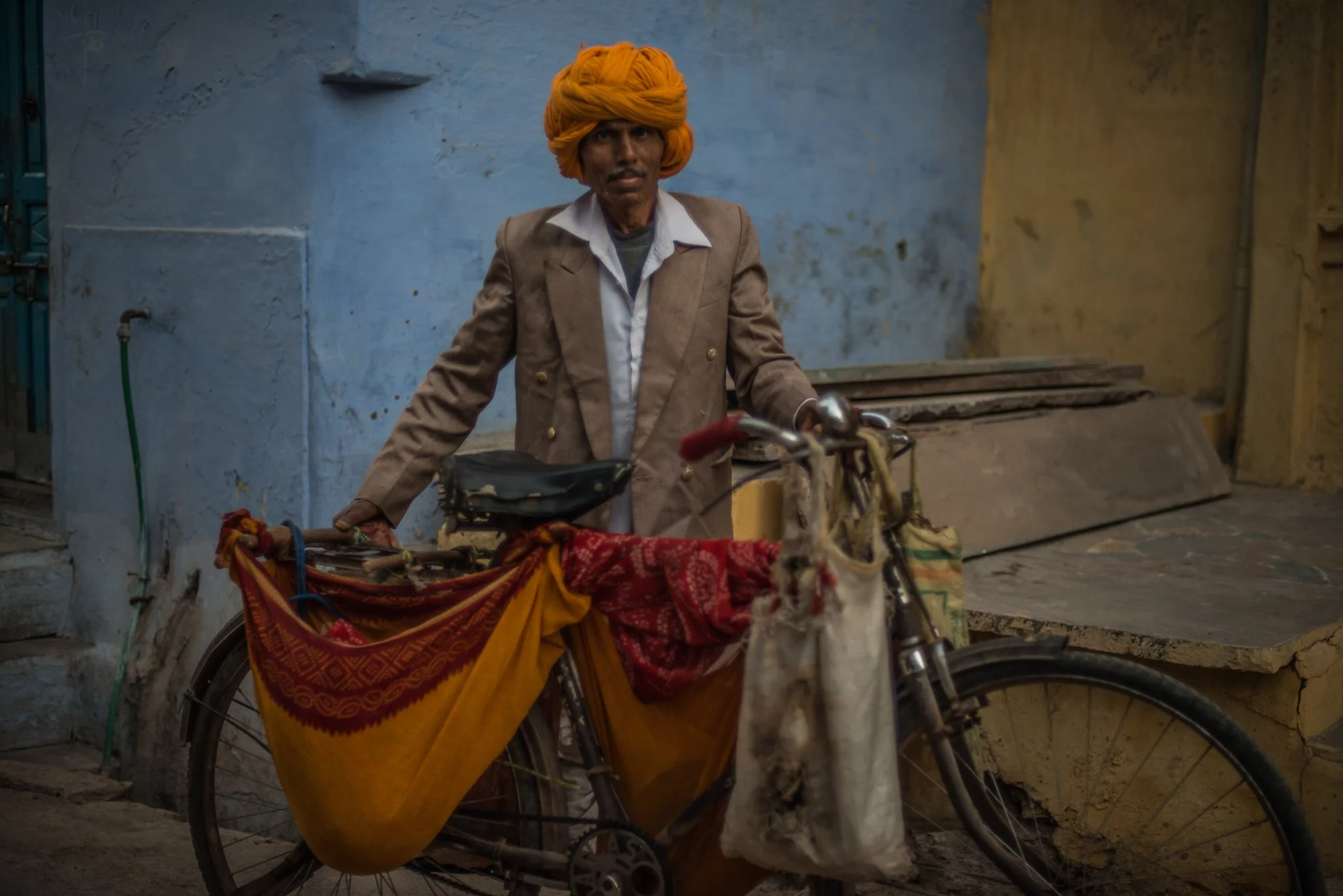 An older man with a yellow turban and a beige blazer standing beside a bicycle with cloth and bags attached, in a street with blue and yellow walls.