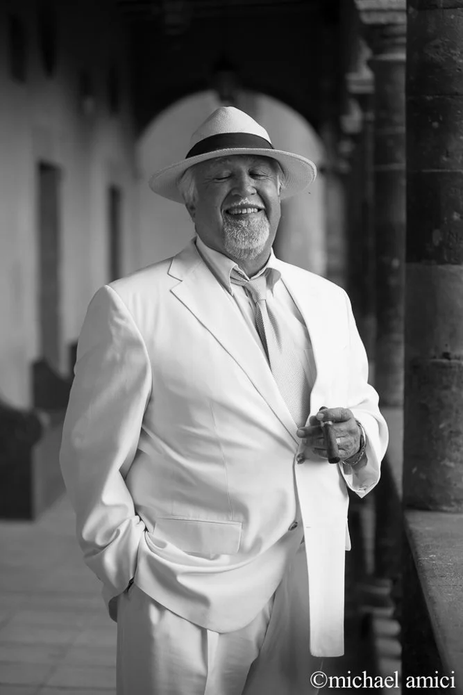 A smiling man in a white suit, wearing a fedora hat and holding a cigar, standing outdoors with arches and stone columns in the background.