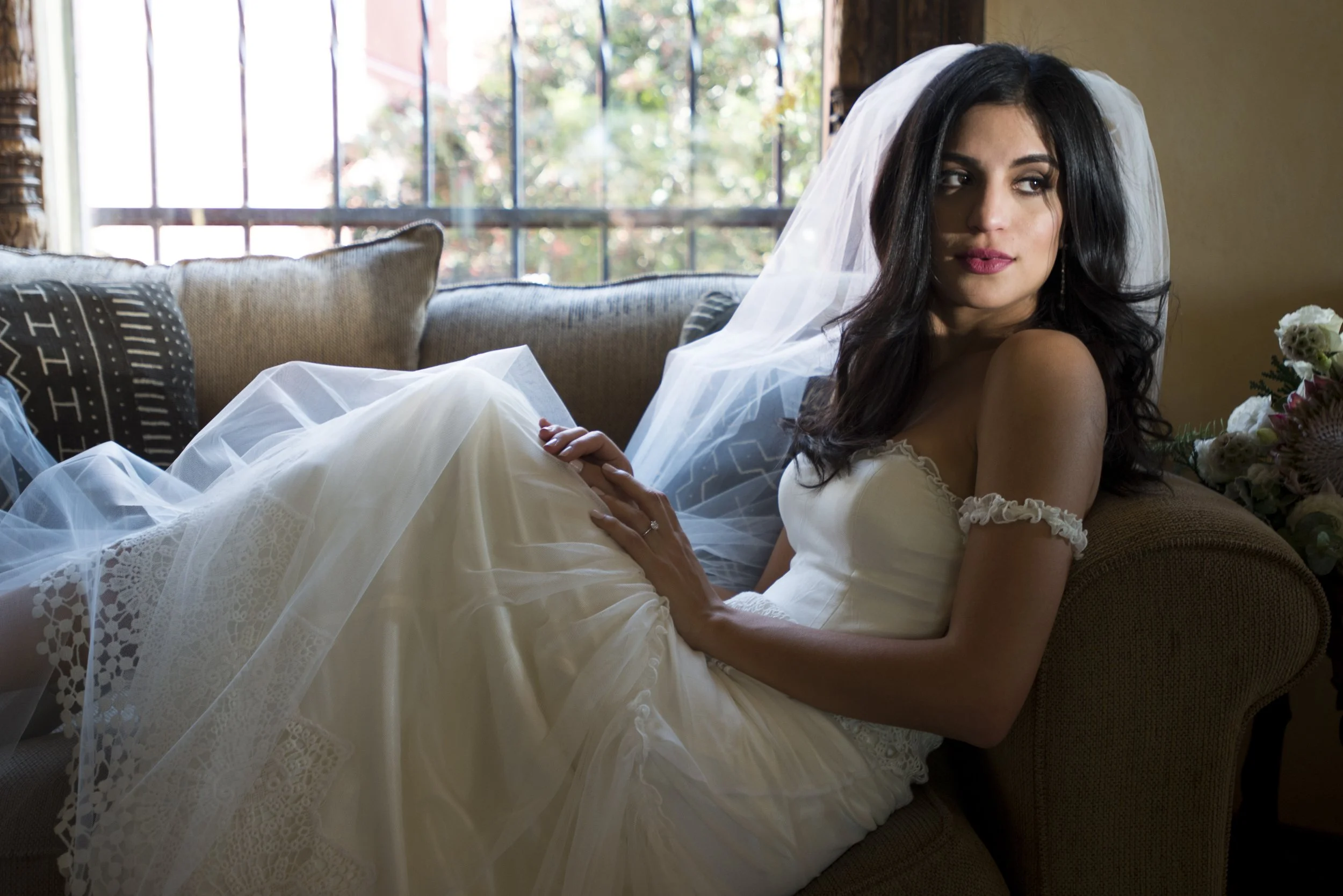 A woman in a wedding dress relaxing on a sofa with a white veil, sitting near a window with colorful outdoor scenery.