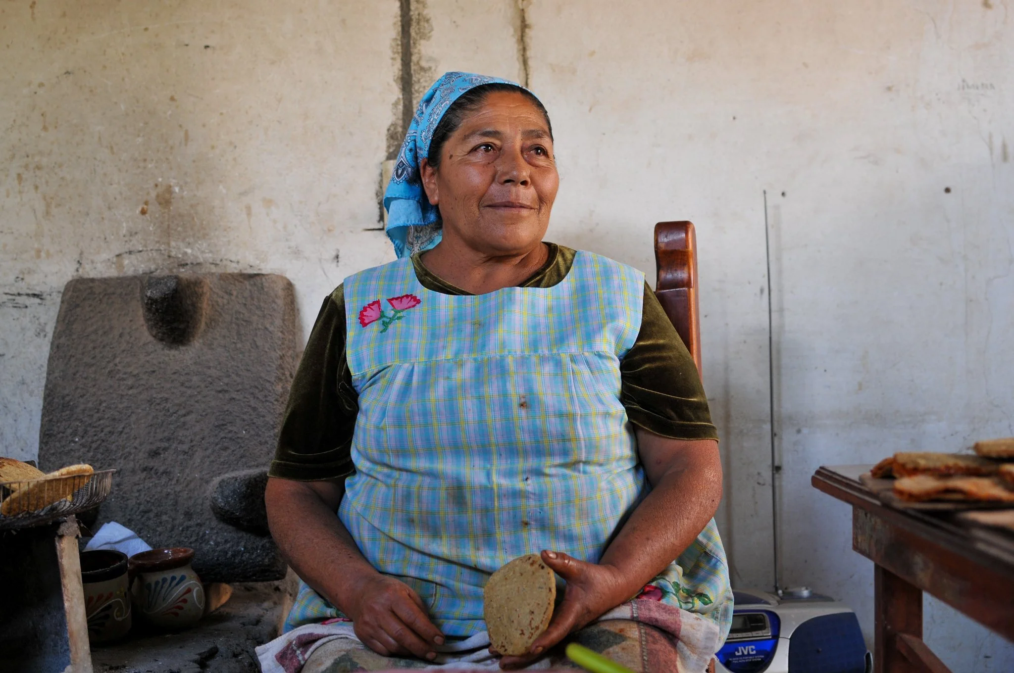 An elderly woman wearing a blue headscarf and a colorful apron sits indoors, holding a round piece of bread.