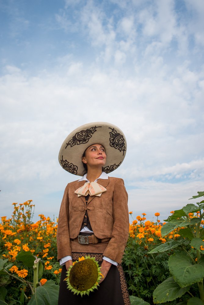 A woman in a large sombrero hat and traditional clothing holding a sunflower standing in a field of orange marigolds, with a mostly cloudy blue sky overhead.