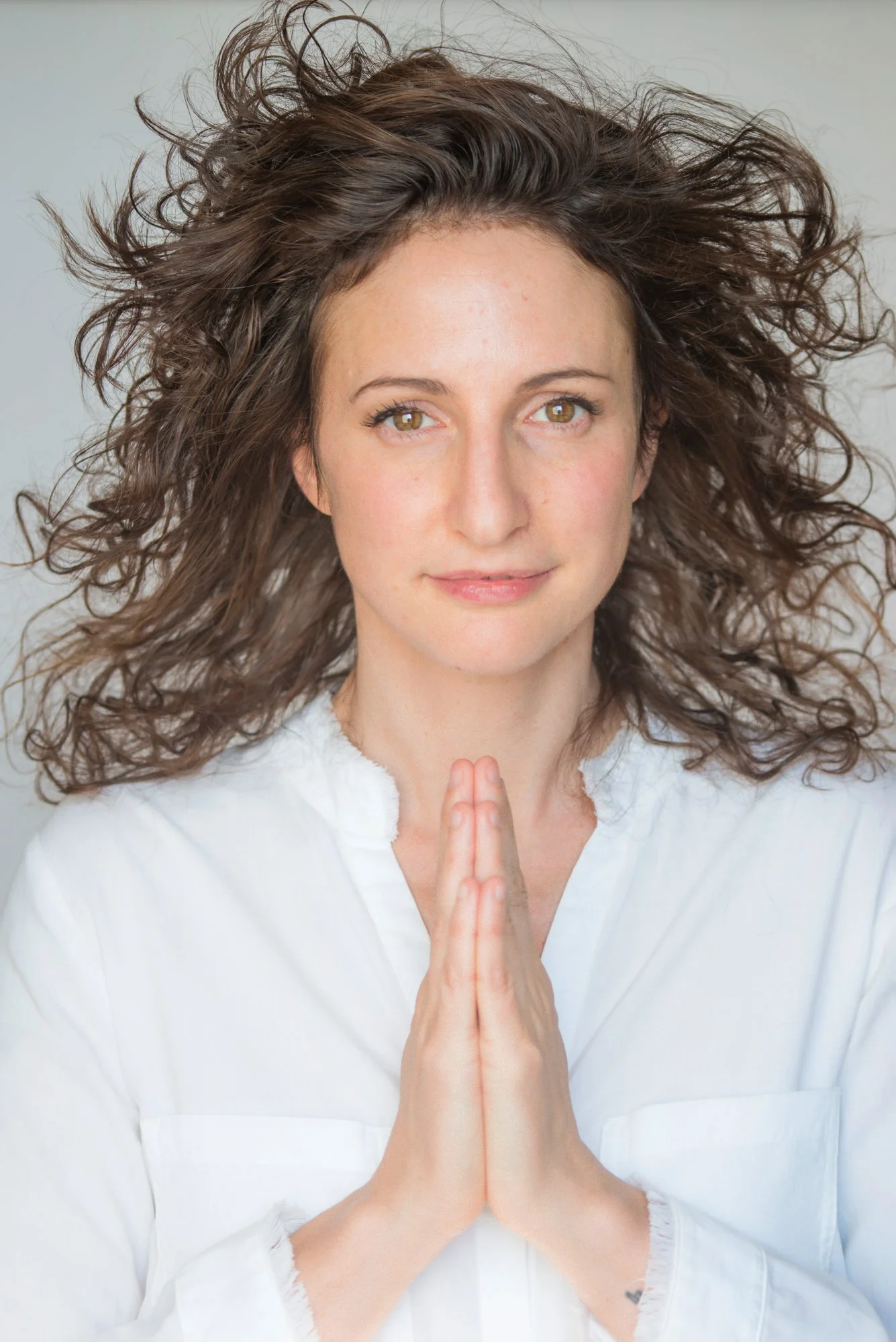 A woman with curly brown hair and hazel eyes holding her hands together in a prayer position, wearing a white top, against a neutral background.