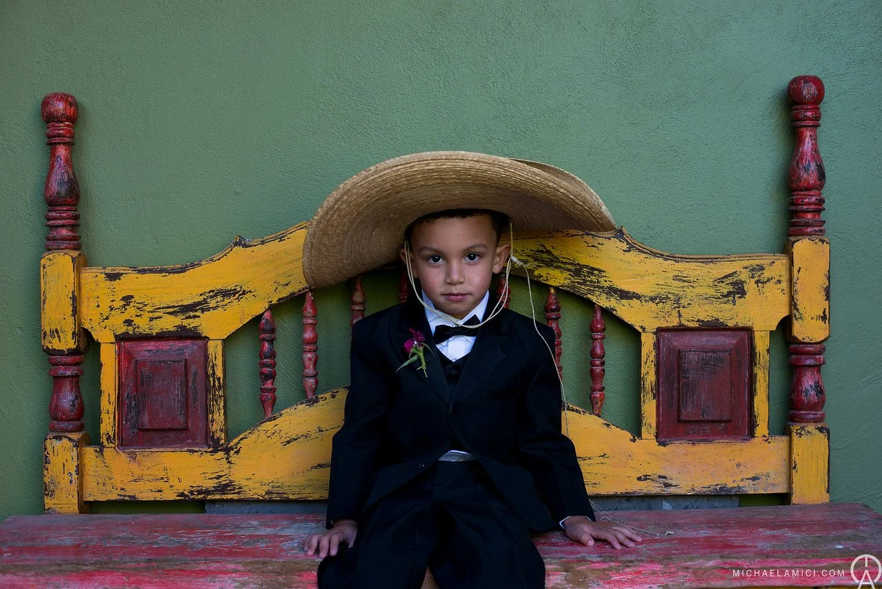 A young boy dressed in a black suit, white shirt, and bow tie, sitting on an old, weathered yellow and red wooden bench against a green wall. He is wearing a wide-brimmed straw hat tilted over his head and has a pink flower pinned to his suit. The bo
