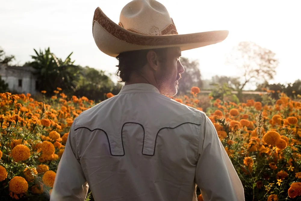 A cowboy standing in a field of orange marigold flowers with trees and a building in the background, during sunset in Mexico. 