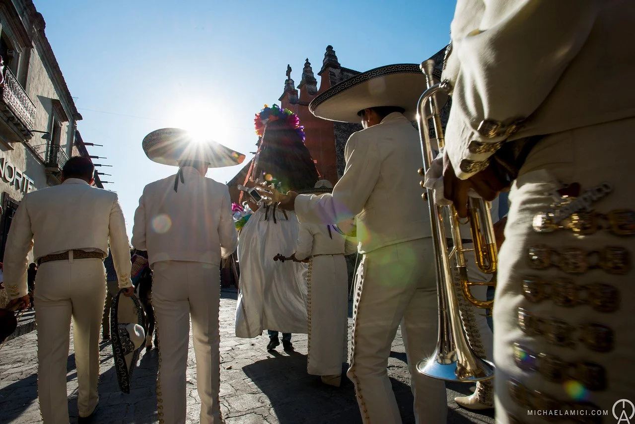 Mexican mariachi band dressed in white suits playing instruments outdoors on a sunny day.