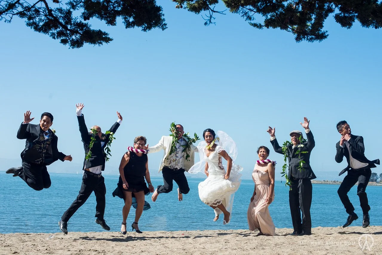 Group of people in wedding attire jumping on the beach near the water, with trees and a clear blue sky in the background.