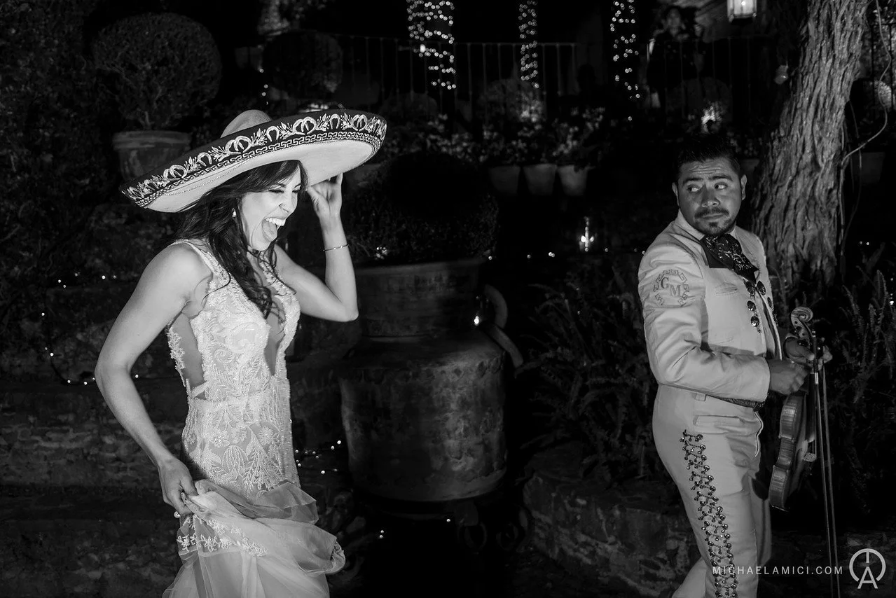 A woman wearing a large sombrero and a lace dress laughing, and a man holding a violin, standing outdoors at night with string lights and a large tree in the background.