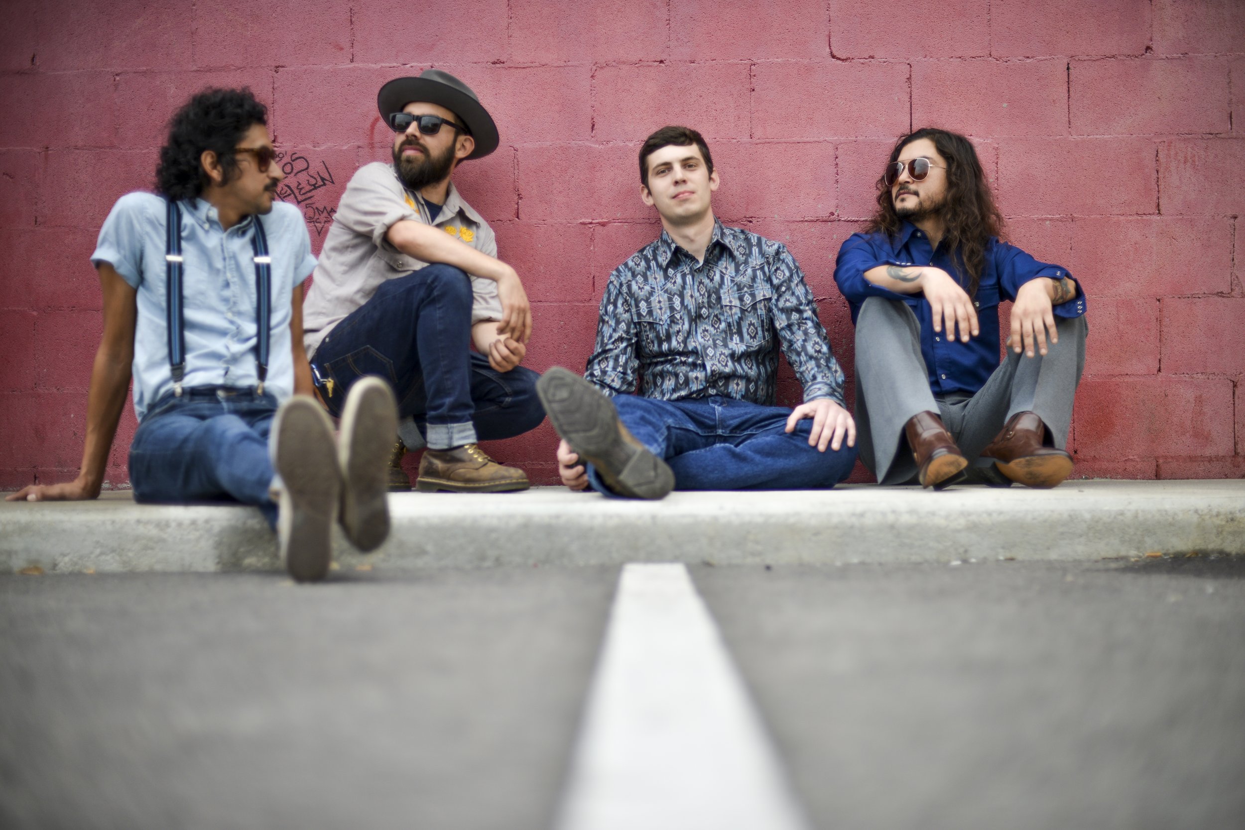 Four men sitting on the curb against a pink wall, one with sunglasses and a hat, another with sunglasses and long hair, one in a button-up shirt, and another in suspenders and a light blue shirt.