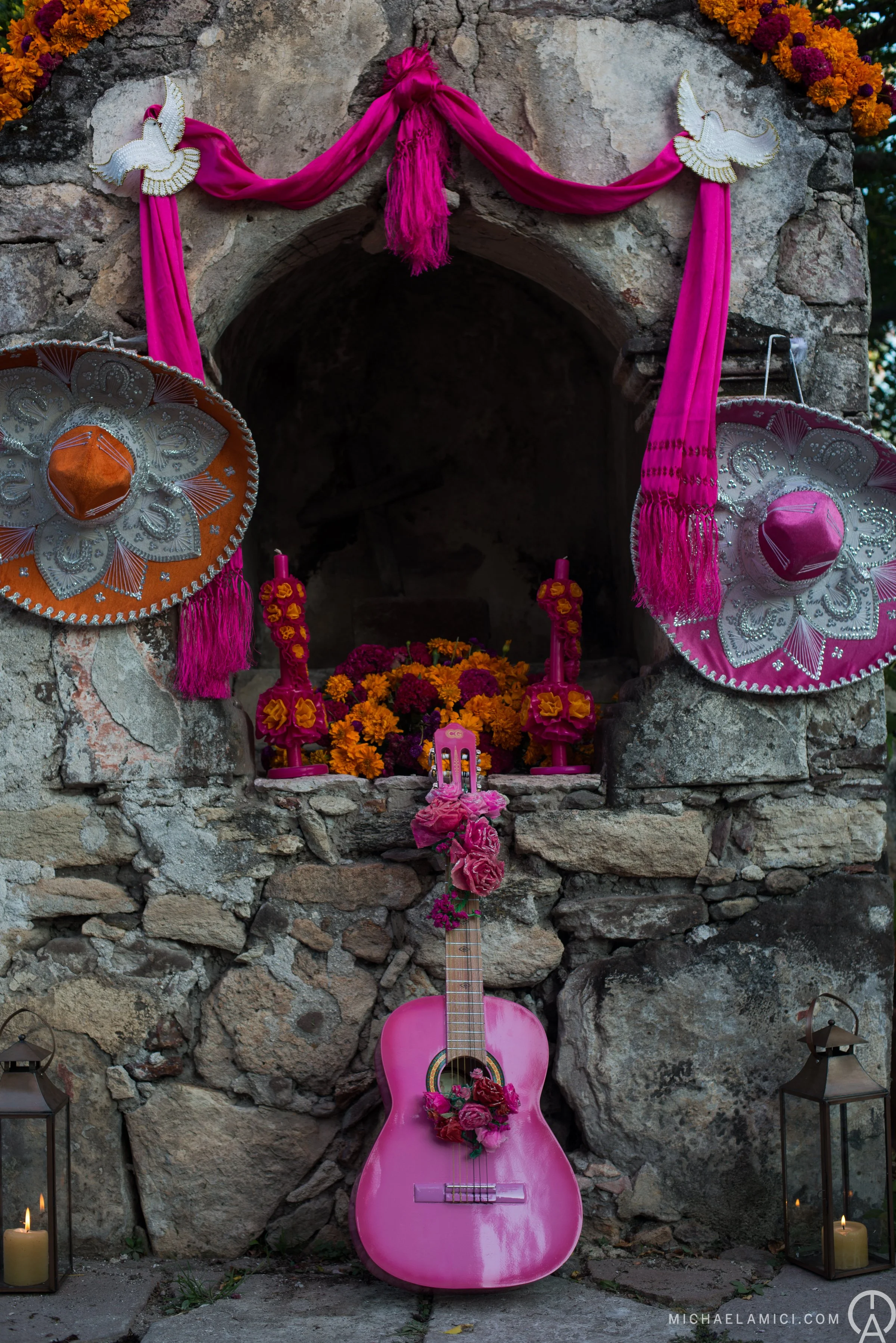 A decorated shrine with a pink guitar, flower arrangements, papel picado, and sombreros, likely for Día de los Muertos festivities.