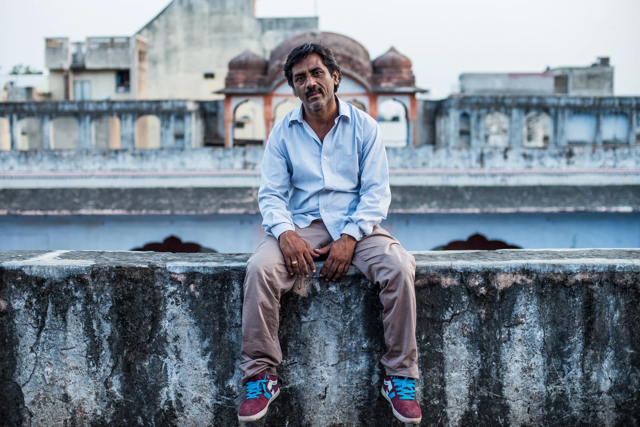 A man sitting on a rooftop wall wearing a light blue shirt, beige pants, and blue and burgundy sneakers, with a cityscape and historic building in the background.