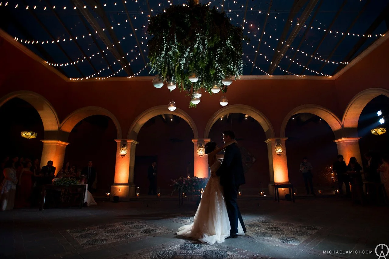 A bride and groom sharing their first dance at a wedding reception, illuminated by warm lighting and string lights hanging from the ceiling.