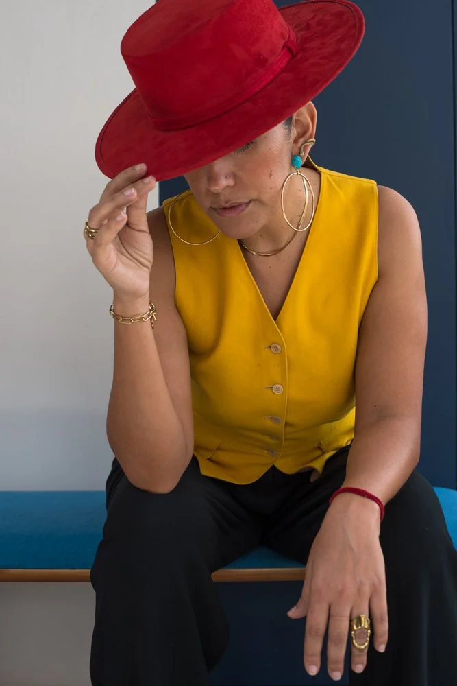 Woman wearing a large red hat, yellow sleeveless top, black pants, and various jewelry, sitting on a blue bench against a dual-colored background, with her face partially hidden by the hat.