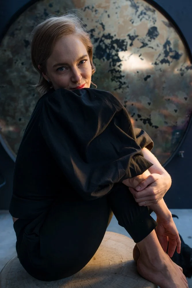 A woman with short blonde hair and fair skin, wearing a black jacket and black pants, sitting on a round wooden surface with her knees drawn up, outdoors with trees and a circular window in the background during sunset.