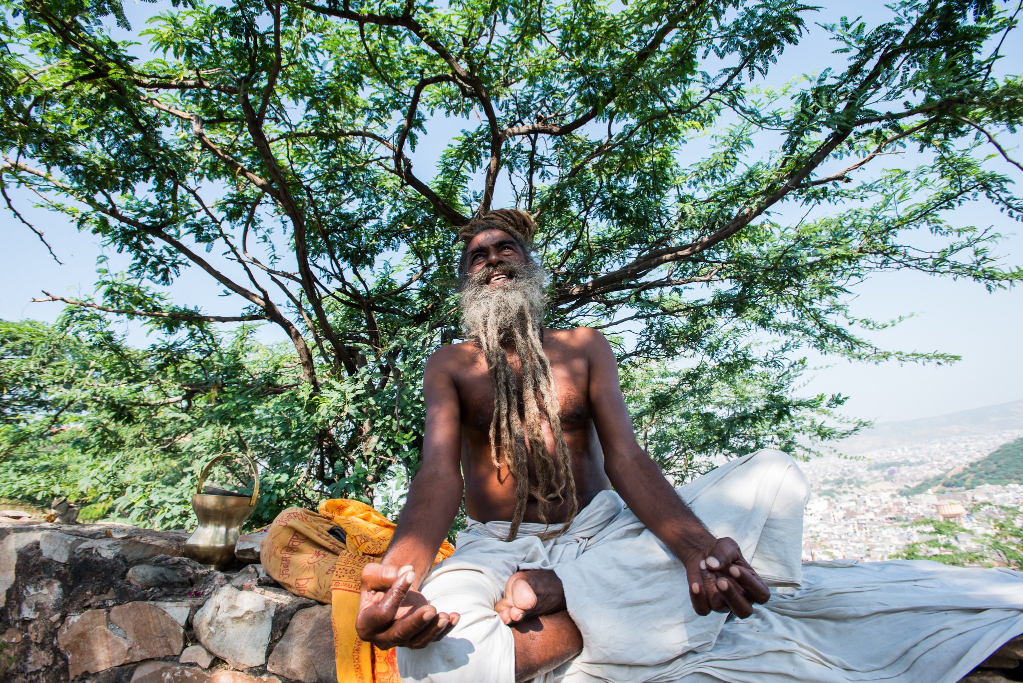 A shirtless man with a long gray beard and dreadlocks meditating outdoors under a large tree, sitting cross-legged on a cloth, with a cityscape in the background.