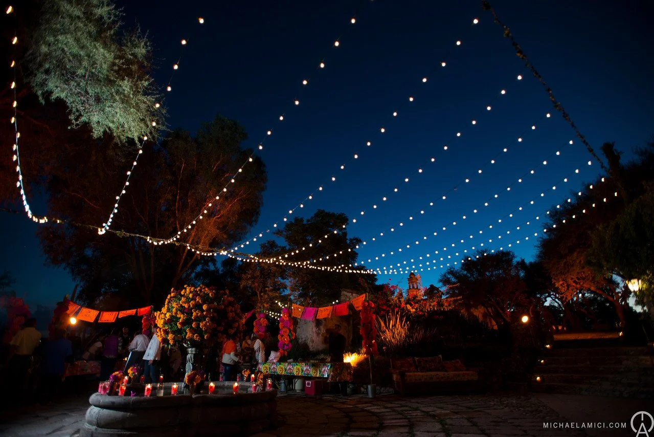 Nighttime outdoor celebration with string lights overhead, colorful flower decorations, and people gathered around with candles and altars.