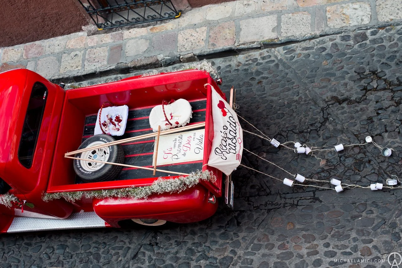 Red miniature truck decorated for Christmas with a small tire, a white towel, and a heart-shaped ornament in the truck bed. A string of small white holiday lights extends from the back of the truck, and the truck is parked on a stone street.