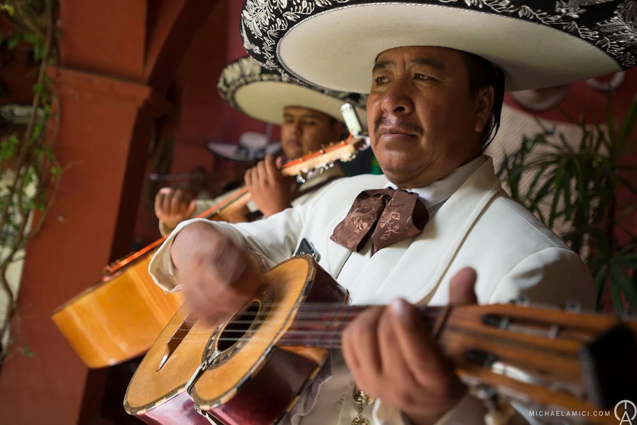 Close-up of a mariachi musician wearing a traditional sombrero and white suit playing a guitar, with another musician in similar attire playing a guitar in the background.