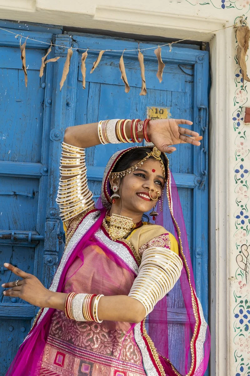 A woman in traditional Indian attire dancing in front of a blue wooden door, wearing vibrant pink, gold, and purple clothing, with numerous gold and red bangles, jewelry, and a bindi on her forehead.