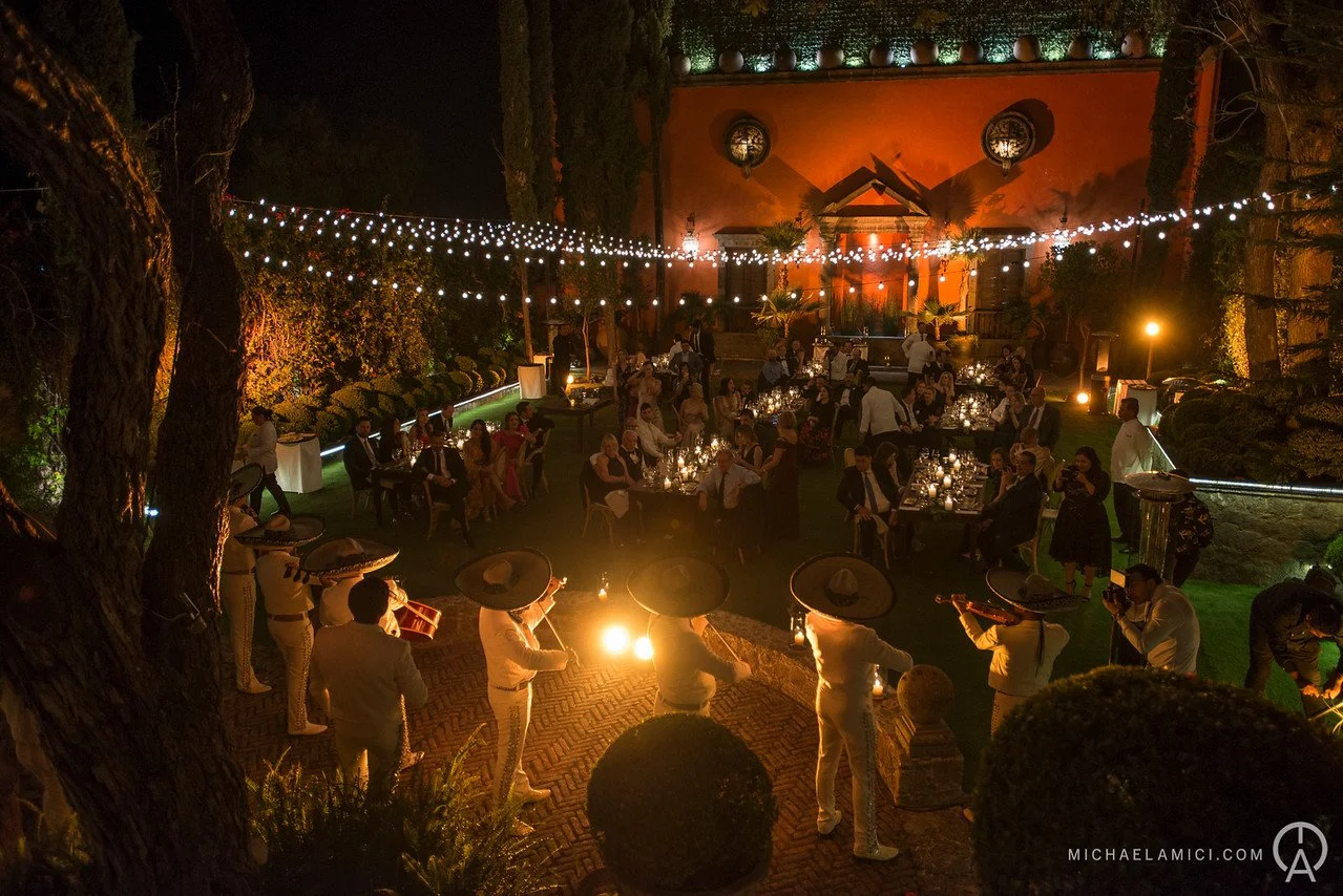 A nighttime outdoor celebration with tables of people dining and a mariachi band playing music. String lights are hung overhead, and a red wall with decorative plants and circular windows is in the background.