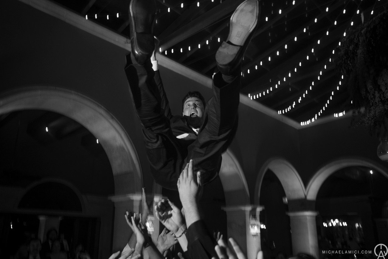 A man in a tuxedo being lifted by a crowd at a celebration or event, with string lights hanging from the ceiling.