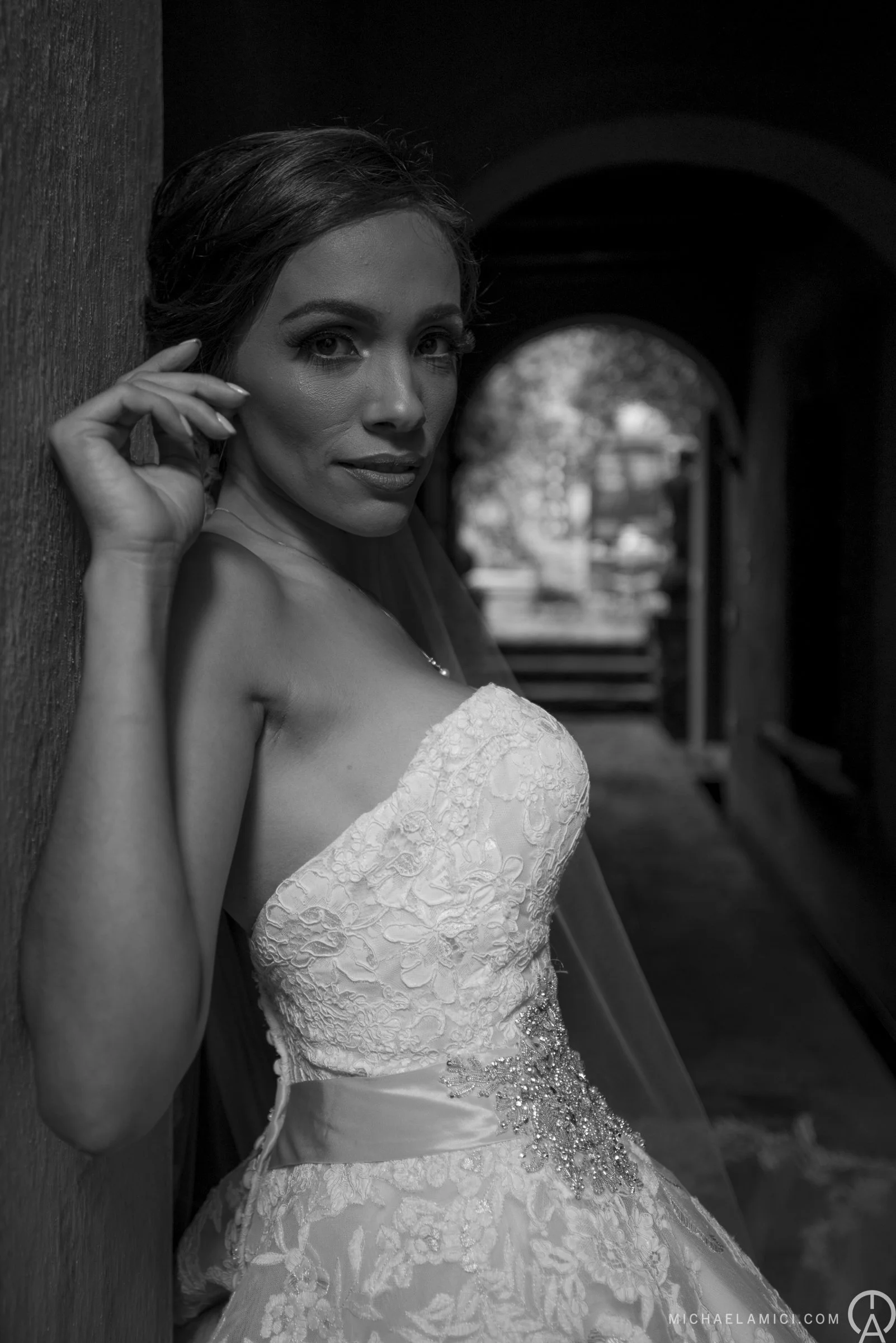 Black and white photo of a woman in a strapless wedding dress, leaning against a wall in a dark corridor, with an archway and trees visible in the background.