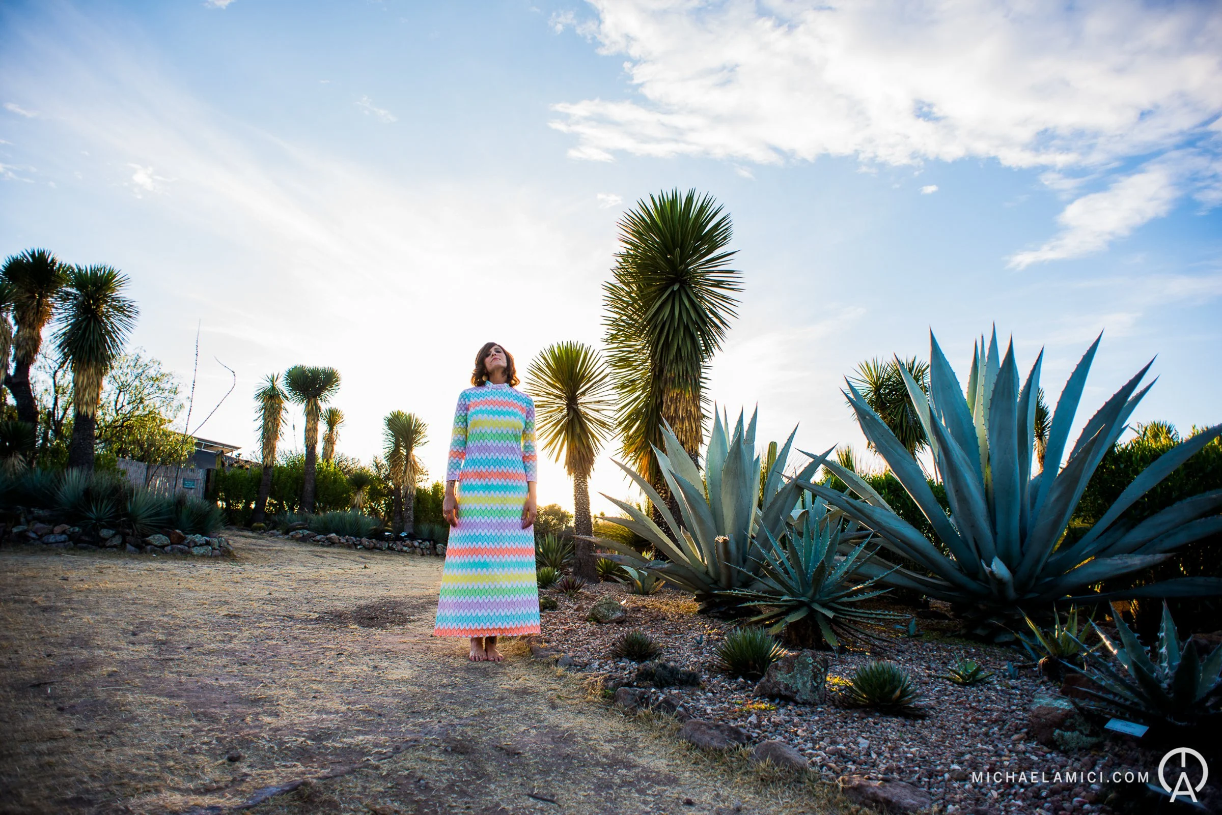 A woman in a colorful, long, zigzag-patterned dress stands barefoot outdoors among desert plants, including large agave and palm trees, under a partly cloudy sky during sunset.