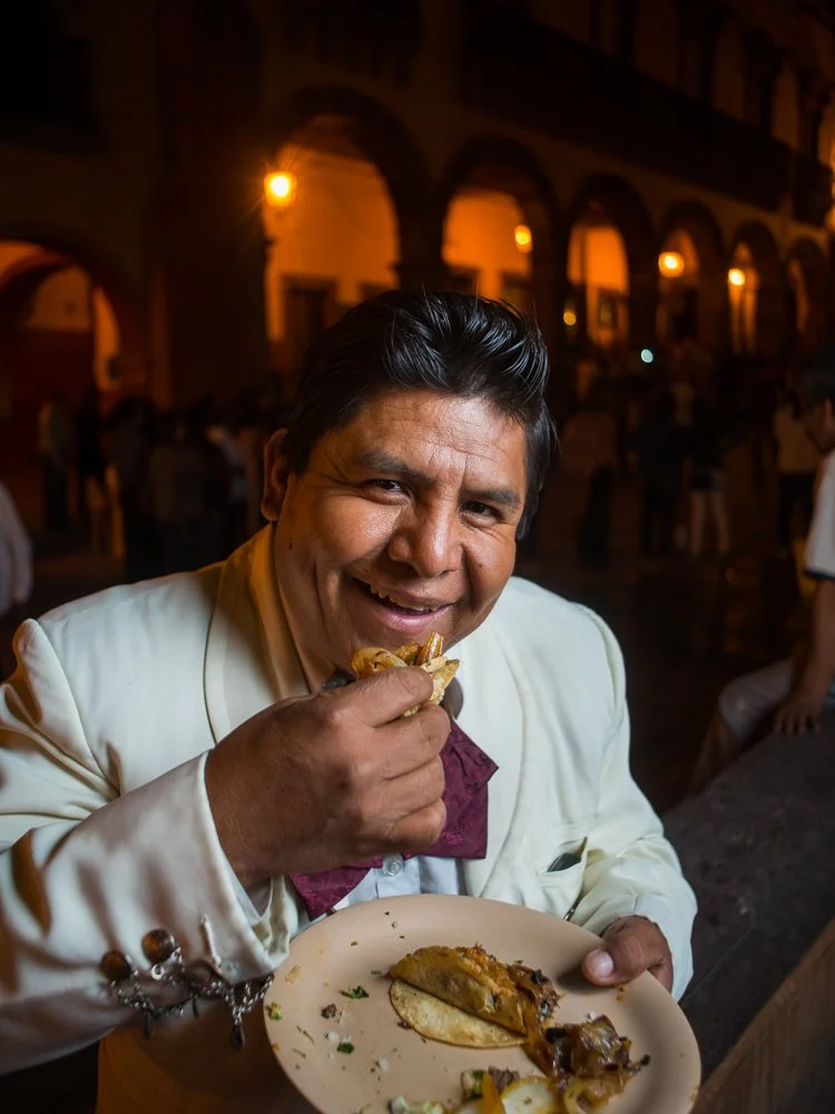 Mariacchi in a white suit eating tacos in a dimly lit restaurant or bar with arches and warm lights in the background.