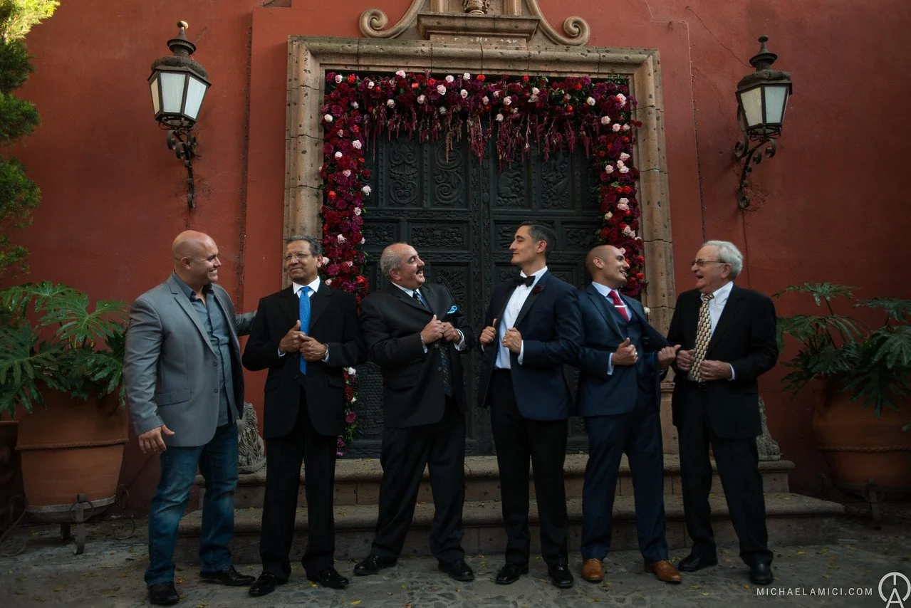 Group of seven men in formal wear stands in front of an ornate, dark door shaped window with floral decorations, outdoors, on cobblestone ground.