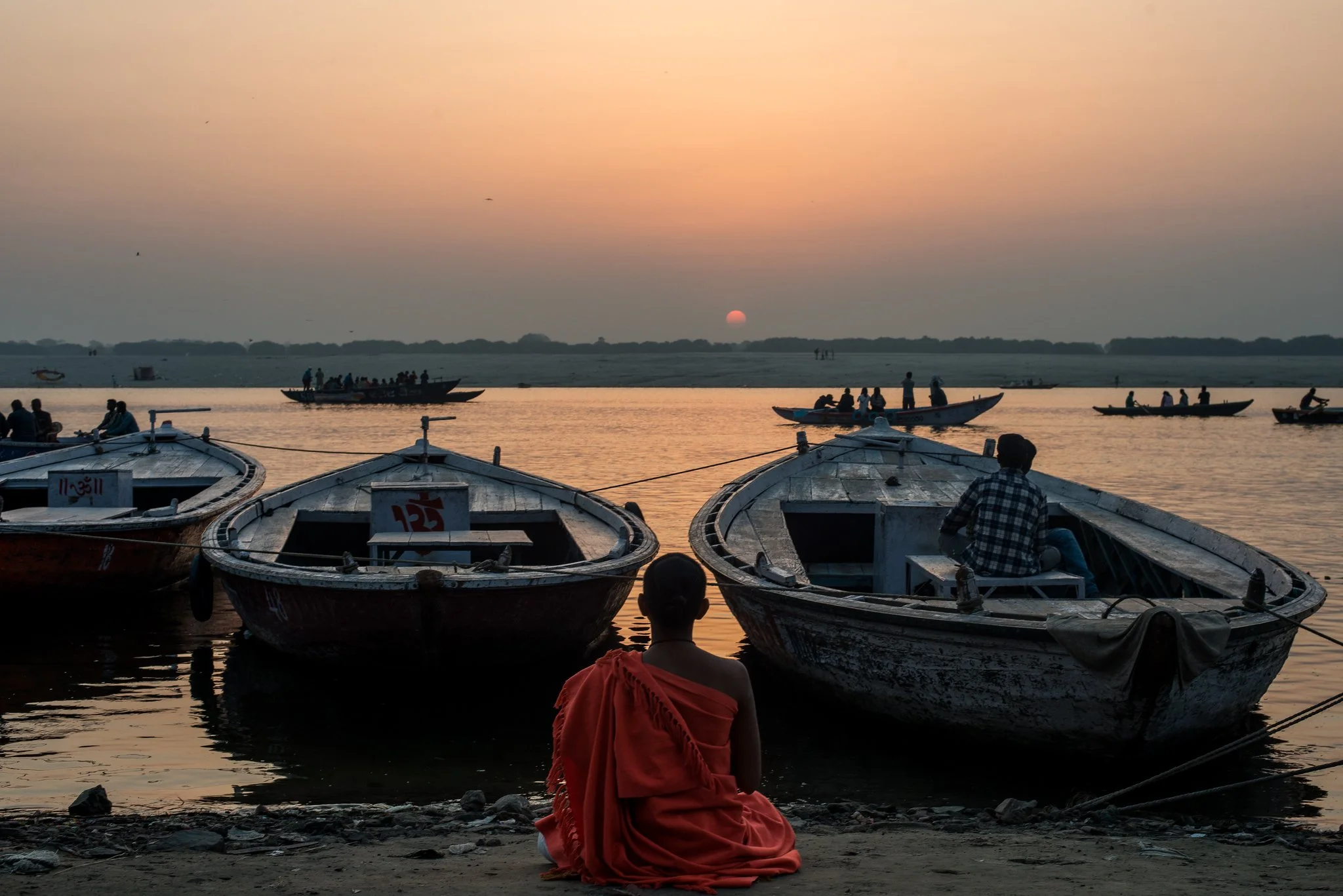 A person dressed in orange sitting on the shore facing a river during sunset, with several boats anchored nearby and others floating on the water, and more boats in the distance.