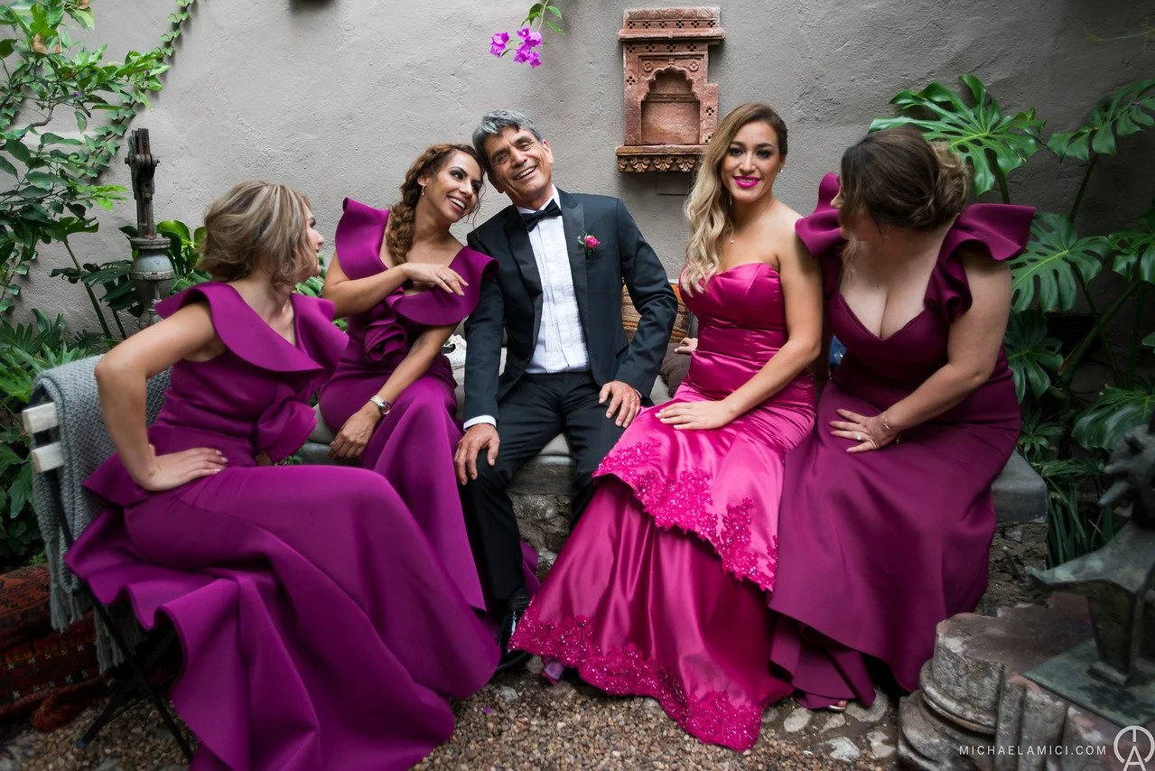 A group of five people, four women and one man, sitting together in a garden-like setting with plants and a stone wall. The women are wearing matching fuchsia dresses with ruffled or embroidered details, and the man is in a black tuxedo with a bow ti