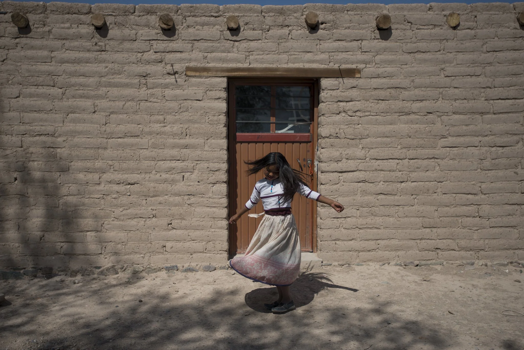 A girl with long dark hair spinning in front of a beige brick wall with a small wooden door, casting a shadow on the dirt ground.